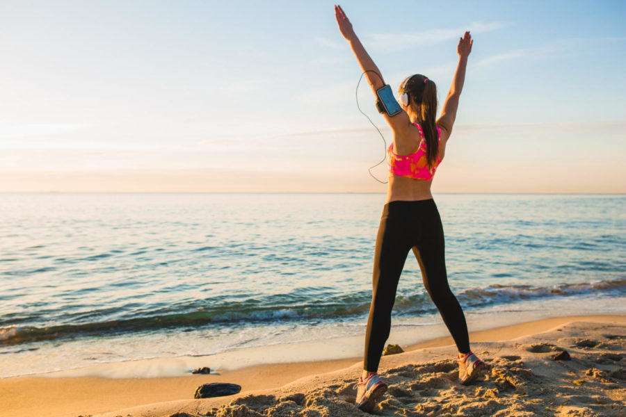 young woman doing sport exercises on sunrise beach in morning, stretching, healthy lifestyle, listening to music on headphones